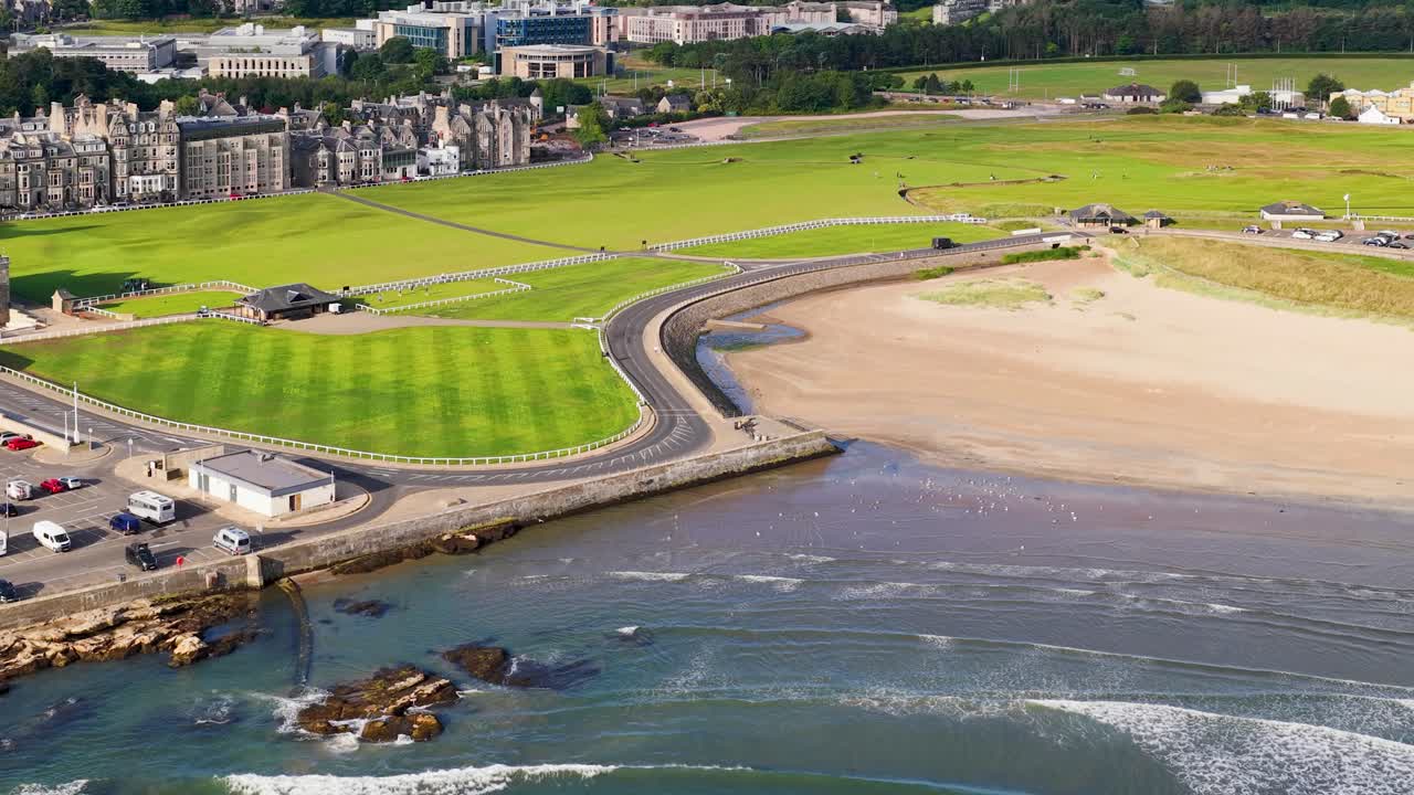 Drone glides above historic coastal golf course, clubhouse, parking, and shoreline under bright daylight