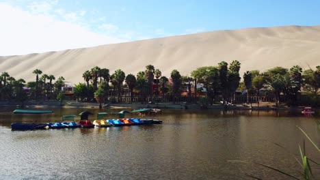 Aéreo,-Tiro-De-Drone,-Hacia-Botes-De-Pedales,-En-La-Laguna-De-Huacachina,-El-Pueblo-Del-Oasis,-En-Un-Día-Soleado,-En-El-Sur-De-Perú