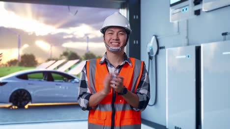 asian male engineer with safety helmet smiling and clapping his hands while standing with home energy storage system in a modern garage