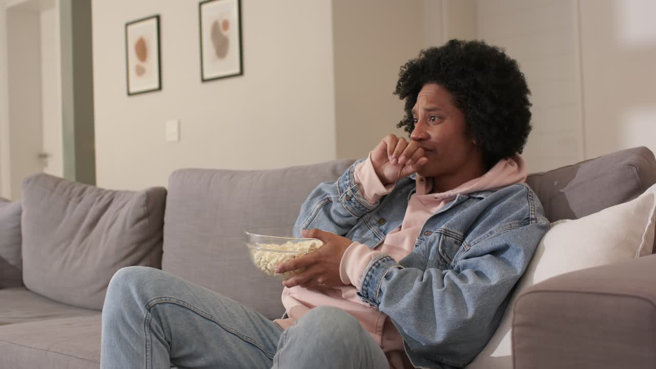 Un uomo di mezza età, percependo una tensione fuori campo, si tocca i capelli, si morde un'unghia e mangia popcorn in salotto.