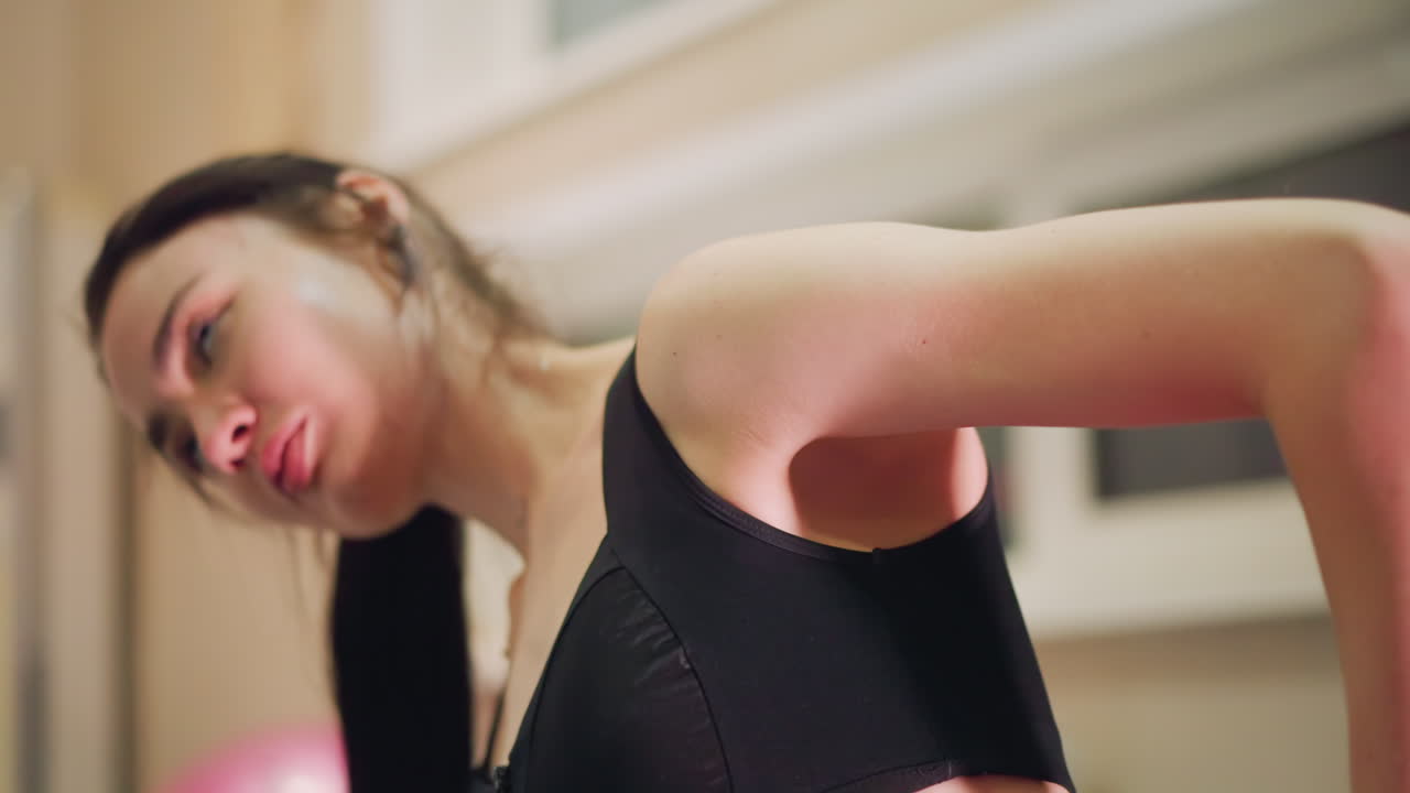 Bailarina caucasiana executando um alongamento lateral em estúdio, vestindo um top esportivo preto. Inclinação controlada do pescoço, sob iluminação suave de fim de tarde. Consciência da respiração, como parte de uma sequência de desaquecimento e recuperação, com uma expressão graciosa.