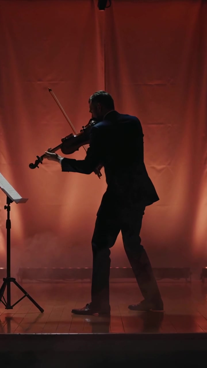 Vertikal video: Violin løftes, mand i mørkt jakkesæt bukker, træder ind på scenen, spiller med nodestativ og mikrofon.