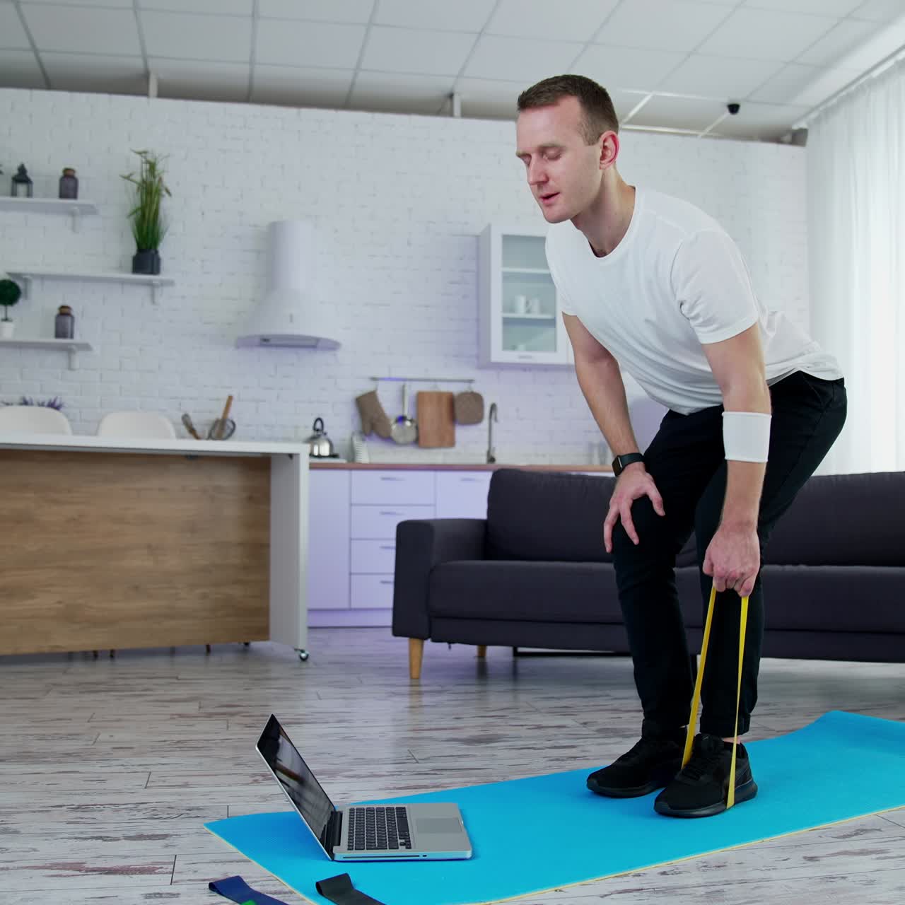 Young man training at home with a sport rubber band. Guy exercising on a mat standing in front of a laptop in kitchen. Healthy lifestyle