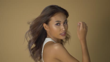 Young-Asian-woman-poses-in-a-professional-fashion-shoot-with-a-fan-blowing-her-hair-as-she-flips-it-back---beige-background