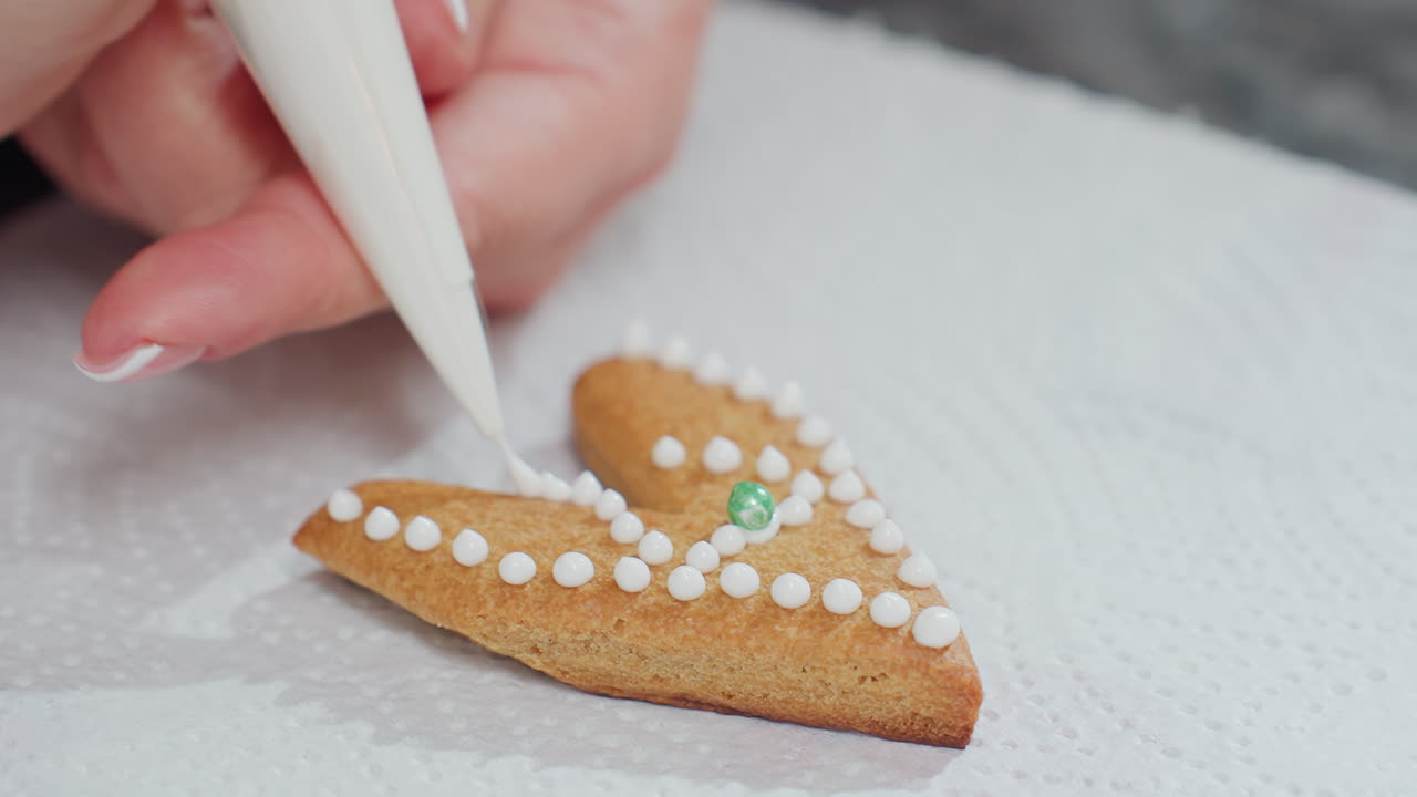 사랑 모양의 쿠키가 색 아이싱 점과 녹색 구슬로 장식되어있는 클로즈업, 색 텍스처 된 종이에 놓여져 있으며, 끔하게 매니큐어 된 손을 가진 손은 조심스럽게 파이핑 가방으로 아이싱을 적용합니다.