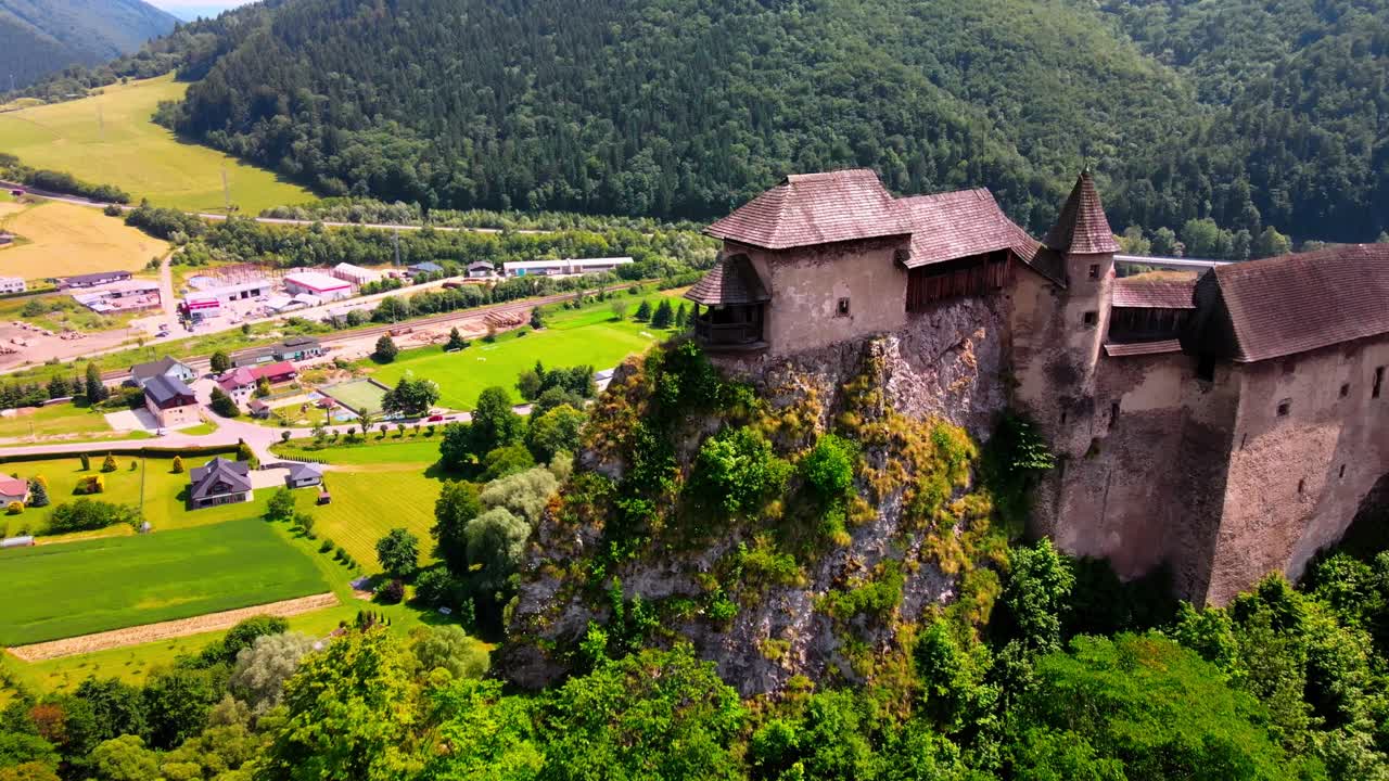 Letecký záběr odhalující středověký Oravský hrad na strmém útesu s výhledem na říční údolí na Slovensku.