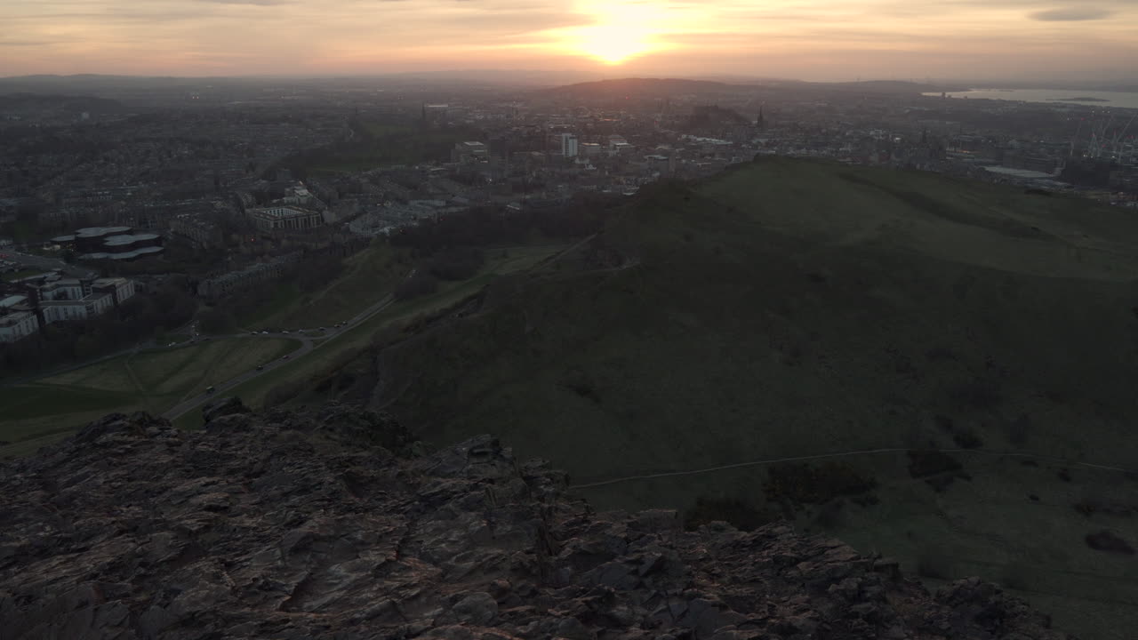 tiro inclinado hacia arriba del paisaje urbano de edimburgo desde la montaña del asiento arturs durante la puesta de sol con colores maravillosos y luz de la hora dorada con autos que pasan por debajo