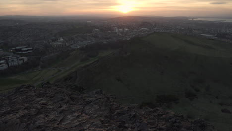 Tilt-up-shot-of-Edinburgh-cityscape-from-the-Arturs-seat-mountain-during-sunset-with-wonderful-colors-and-golden-hour-light-with-cars-passing-by-down-below