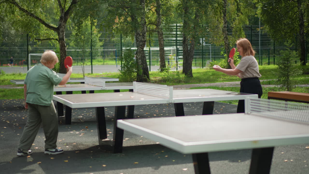 Park Table Tennis White Woman Boy Enjoying Friendly Outdoor Rally Under Leafy Trees, Red Paddles, Concrete Tables, Summer Sunlight, Smiles, Casual Clothing, Playful Competition, Motion And Focus
