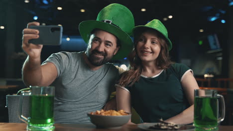 Joven-Y-Mujer-Con-Sombreros-Irlandeses-Con-Jarras-De-Cerveza-Verde-Tomando-Un-Selfie