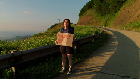 Alleinreisende-Asiatische-Reisende-Trampt-Mit-Einem-Schild-Mit-Der-Aufschrift-„Marry-Please“-Am-Straßenrand-Einer-Bergstraße-Während-Eines-Roadtrip-Sommerurlaubs-Rucksack-Abenteuers