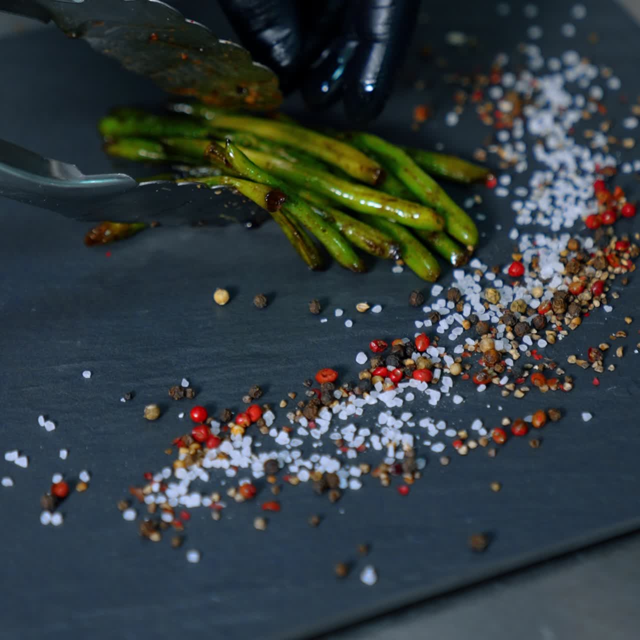 Black charcuterie board with salt and peppers on. Hands in black gloves use tongs to lay the fried beans on the board. Vertical screen. Close up