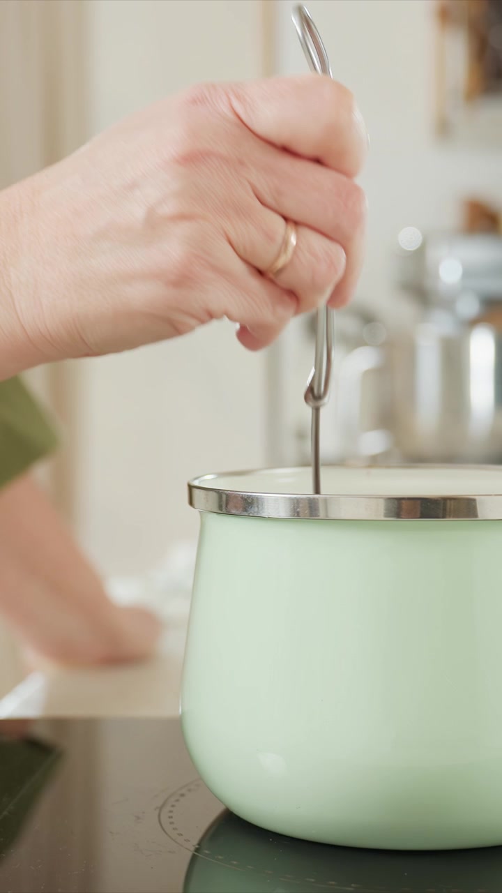 Hand with gold ring repeatedly lifts and lowers metal hook. Background shows blurred kitchen elements. Close up view focuses on precise interaction between human hands cooking