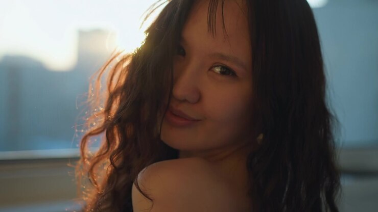 Primo piano di una donna con lunghi capelli mossi vista di spalle e di profilo, mentre la luce del sole filtra attraverso i vetri della finestra rivelando un paesaggio urbano leggermente sfocato. La scena cattura la consistenza dei suoi capelli sciolti