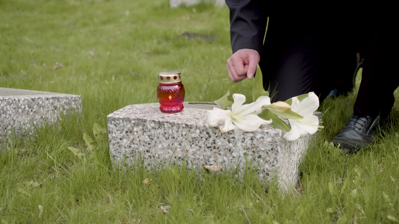 검은 양복을 입은 알아볼 수 없는 남자가 무릎을 꿇고 묘지의 묘비에 꽃과 촛불을 얹고 있다