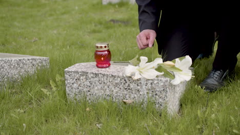 hombre irreconocible con traje negro arrodillado y poniendo flores y una vela en una lápida en un cementerio