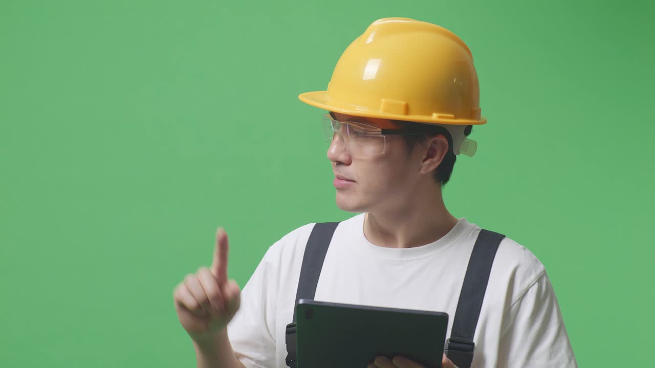 안경과 안전 헬을 착용 한 아시아인 노동자가 태블릿을 사용하고 녹색 화면 배경 스튜디오에 서있는 동안 으로 가리키고 있습니다.
