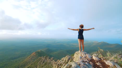 Young-woman-with-arms-raised-at-mountain-peak-overlooking-jungle-landscape