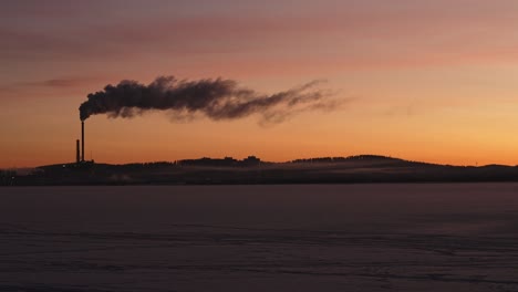 Silhouette-of-smoking-power-plant-in-morning-sun-across-frozen-lake