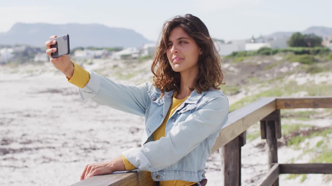 mujer de raza mixta feliz tomando una selfie con un teléfono inteligente y sonriendo en el soleado paseo marítimo junto al mar