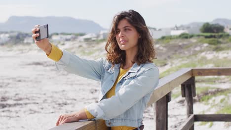 Happy-mixed-race-woman-taking-selfie-with-smartphone-and-smiling-on-sunny-promenade-by-the-sea