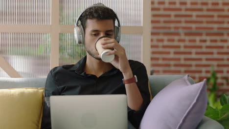 young hispanic man using laptop computer browsing online messages sharing network communication student drinking coffee enjoying listening to music in modern office