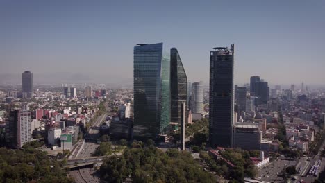 Toma-De-Cámara-Que-Desciende-De-Los-Rascacielos-En-La-Avenida-Paseo-De-La-Reforma-En-La-Ciudad-De-México-En-Un-Día-Soleado-Y-Claro,-Junto-A-Ella-Hay-Una-Gran-Avenida-Con-Varios-Autos-Circulando