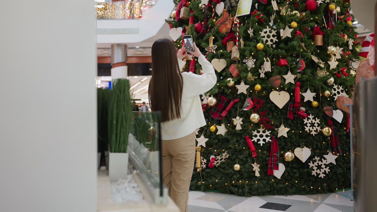 Lady taking picture of beautifully decorated Christmas tree in shopping mall with shoppers seated in restaurant, bokeh light effect in background