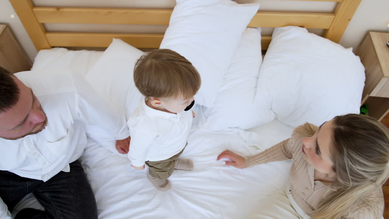 Mom and dad sit on the big bed. Little baby boy with pacifier in mouth stands up looking at parents. Top view.