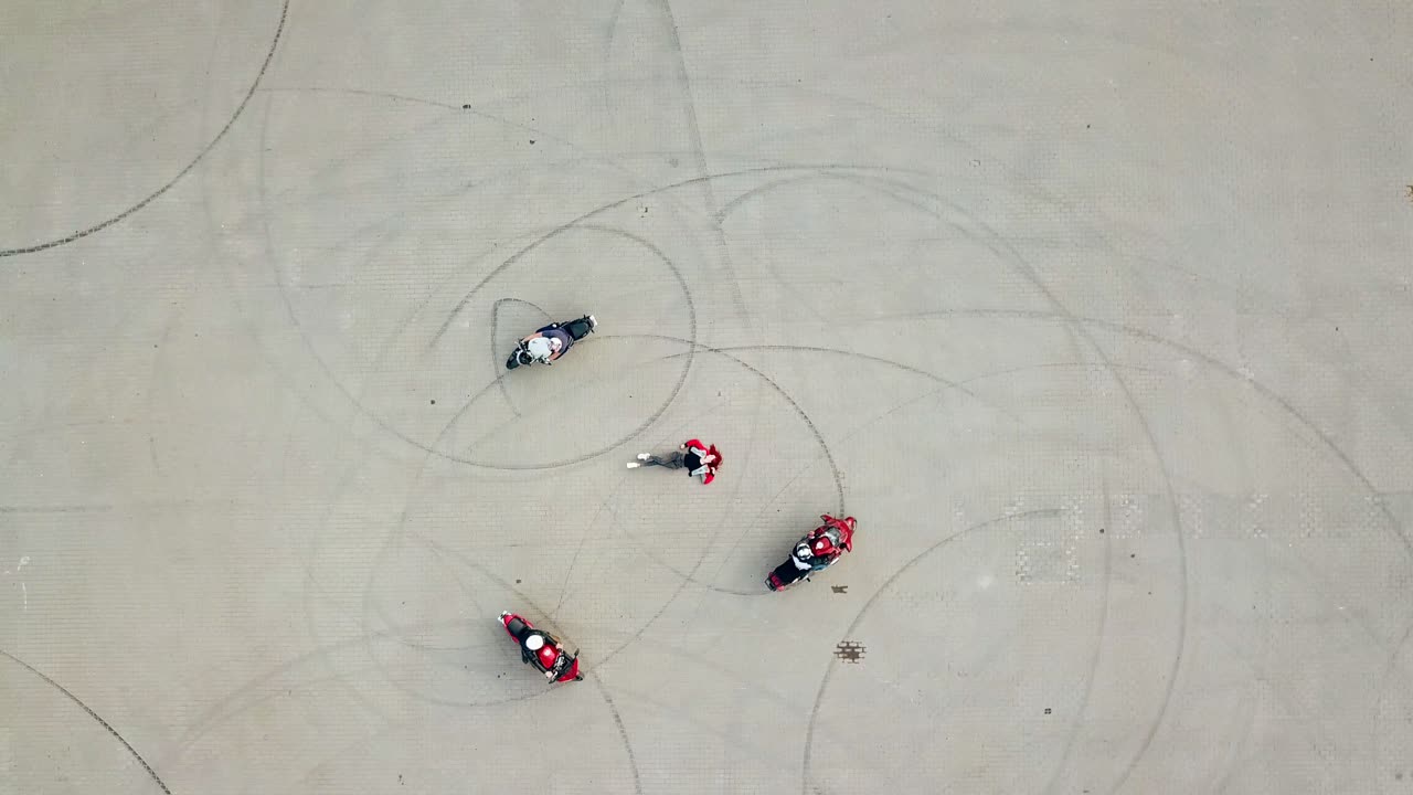 Three professional bikers on their motorcycles riding on a road in the form of a circle. Smiling beautiful young woman lying in the centre of the pavement. Camera moves top down.