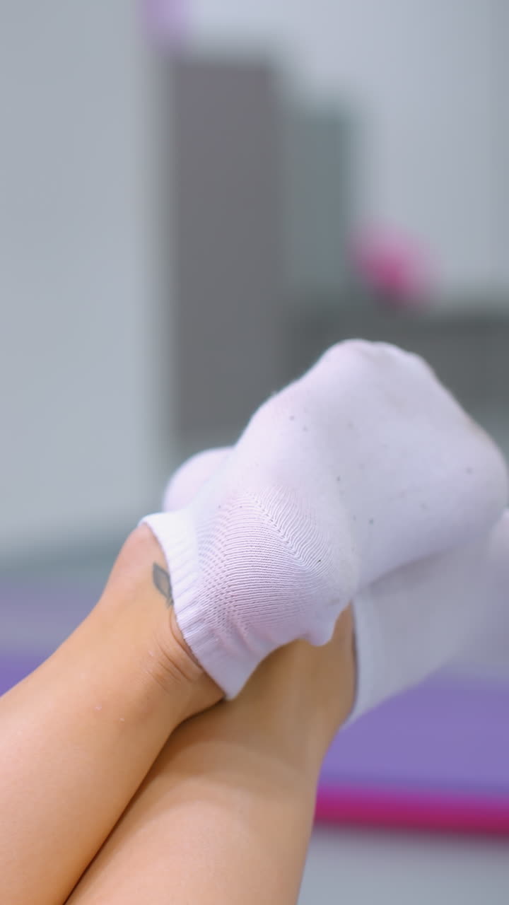 primer plano de piernas con calcetines blancos cruzados en los tobillos balanceándose hacia adelante y hacia atrás, alfombra de yoga púrpura visible en el fondo, movimiento relajado y casual en un entorno interior