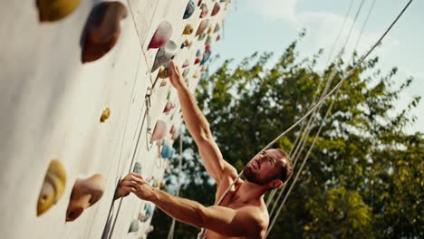 Side view: a sporty guy in black shorts with a sculpted torso climbs up the climbing wall during a rock climbing lesson with belay on a sunny summer day