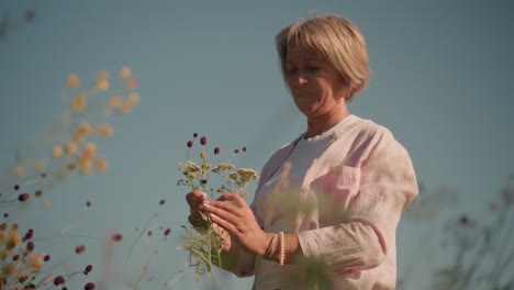 gardener in pink open shirt and white polo arranging flowers in hand under clear blue sky, wearing pink beads on left wrist, with background featuring grassy field and blurred flowers