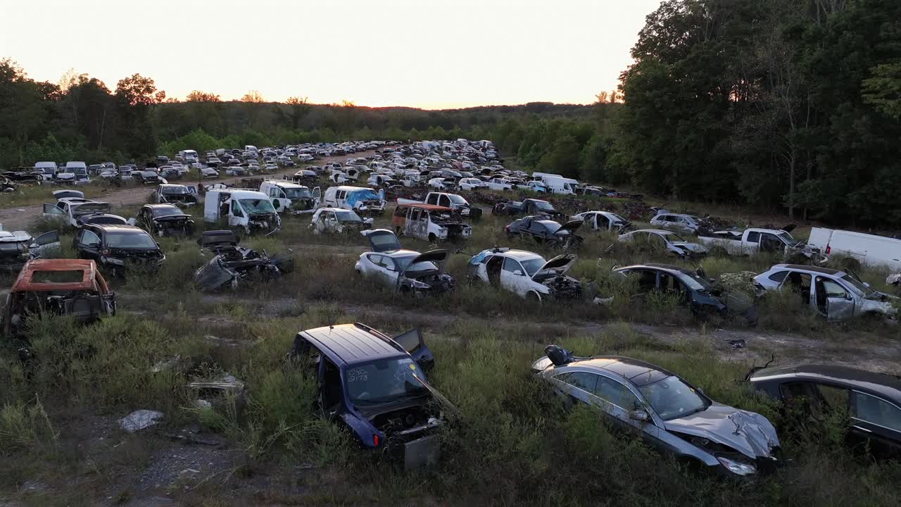 Drone low flight over several rows with destroyed cars and vehicles on junkyard. Sunset time in forest landscape. American