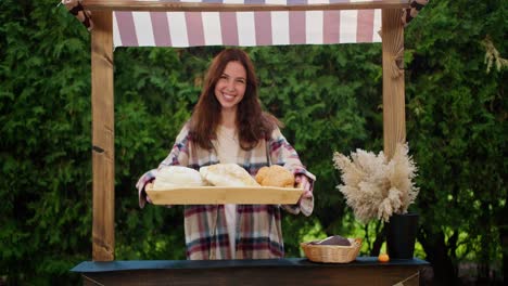 A-happy-brunette-girl-holds-a-tray-with-various-delicious-pastries-in-her-hands-during-her-work-in-a-bread-shop-against-the-backdrop-of-green-coniferous-trees-in-summer