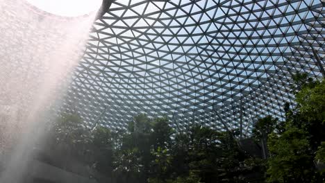 a dramatic indoor waterfall pours from a circular skylight in a geometric glass dome, surrounded by vibrant greenery and natural daylight, with a stationary camera angle