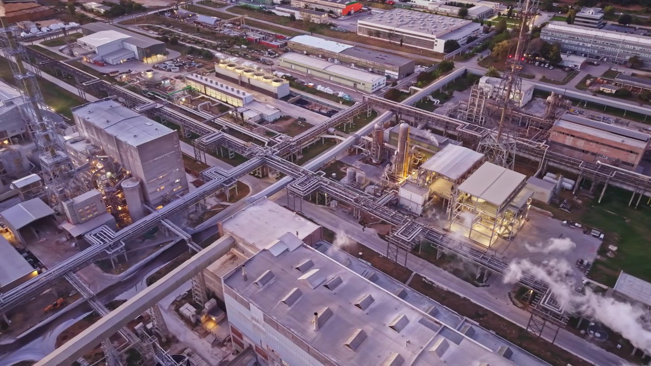Instalação industrial durante a noite com luzes e fumaça no ar, exibindo vários edifícios e infraestrutura