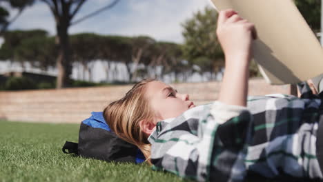 young girl relaxing on grass reading book outdoors, enjoying sunny day, at school