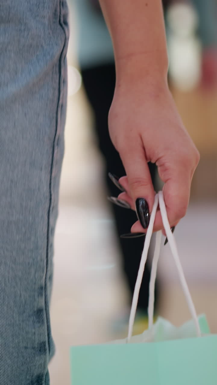 vista trasera de cerca de una mujer con vaqueros y uñas pulidas negras que lleva bolsas de compras rosadas y menta mientras camina en un centro comercial moderno, figuras borrosas en el fondo con reflejos de luz suave en el suelo