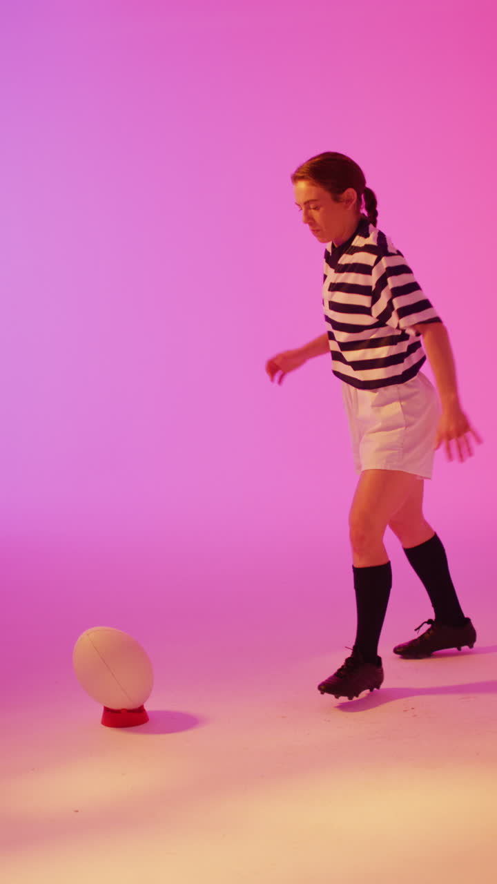 Video vertikal pemain rugbi wanita Kaukasia menendang bola rugbi di bawah pencahayaan neon merah muda
