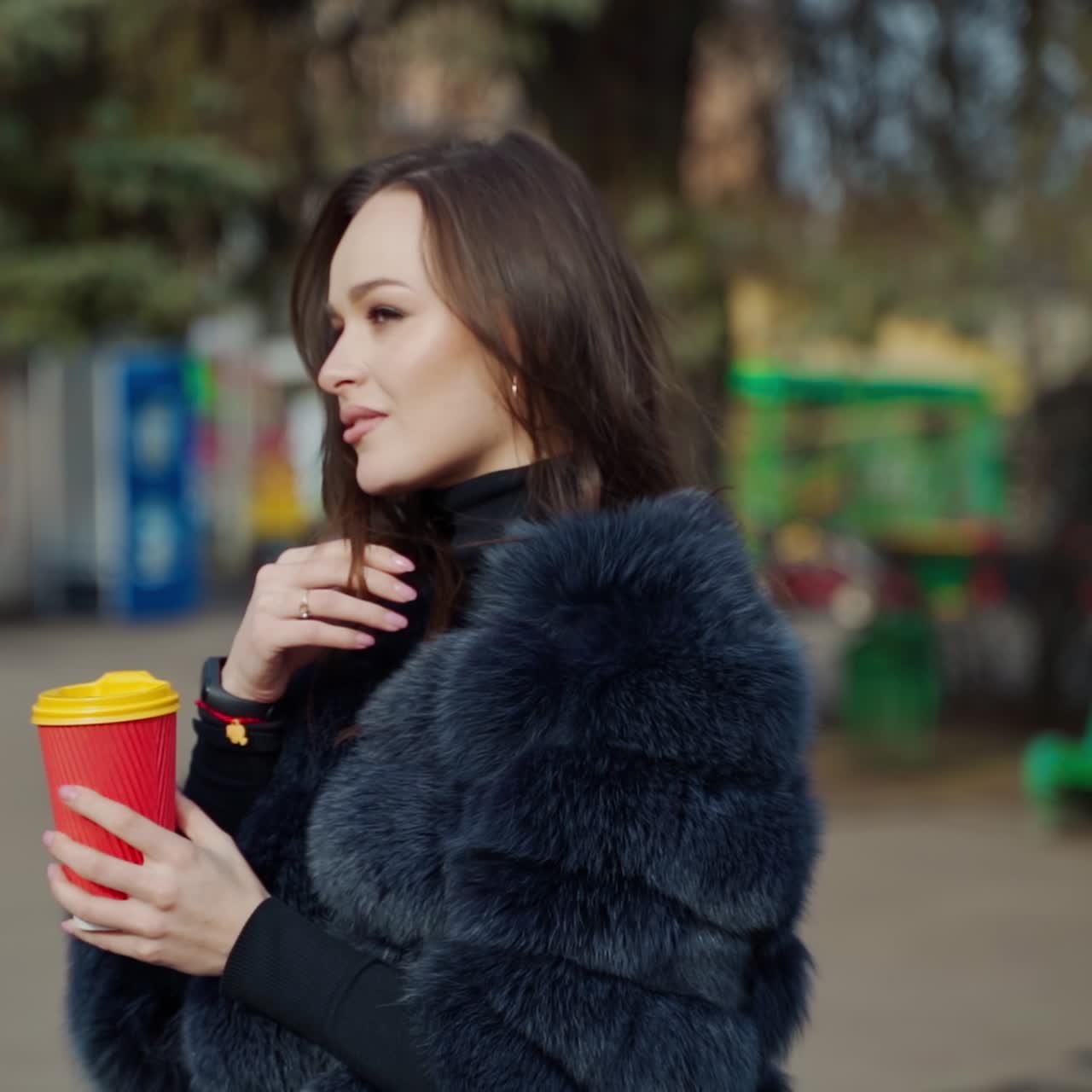 Beautiful woman with a plastic cup of coffee in the park. Portrait of brunette girl in fur coat posing on camera outdoors. Slow motion.