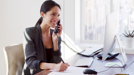 Una-Joven-Asiática-Hablando-Por-Teléfono-Sonriendo-En-Su-Escritorio-De-Oficina