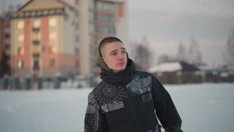 teenager gliding on ice surface with soft blur background featuring building, wearing patterned winter jacket and hoodie, expression looking sideways, snowy urban ice rink setting under sky