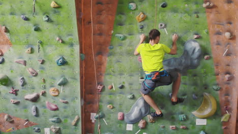 Male-climber-climbs-the-cliff-wall-in-the-room-reaching-and-gripping-hold.-A-professional-concentrated-climber-moves-up-the-wall-the-camera-rises-and-lays-down-his-movements.-Boom-and-truck-follow-camera.