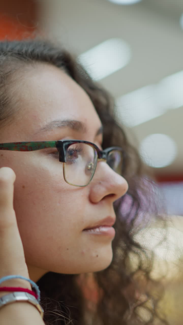 mujer joven con el cabello rizado de pie pensativo en el centro comercial bien iluminado, ajustando su cabello con una expresión suave, el fondo cuenta con una suave iluminación bokeh