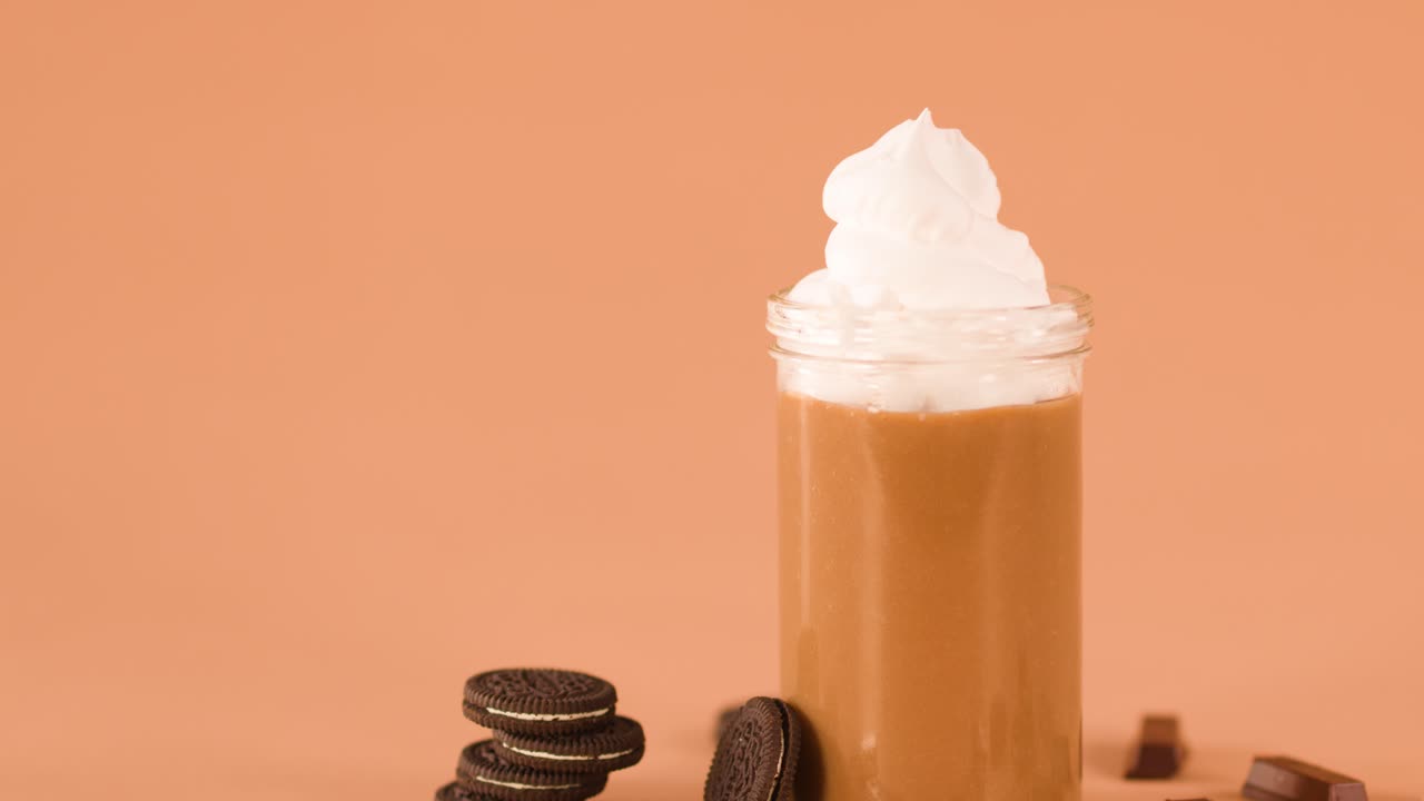 Bir el, etrafı kurabiyelerle çevrili ve yumuşak ışık altında, çikolata barını krem şanti ile kaplanmış milkshake'in içine yerleştiriyor