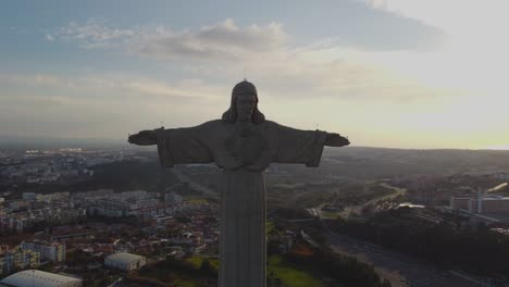 drone hace un círculo completo alrededor de la poderosa estatua de cristo rei jesucristo con una vista espectacular en el horizonte de lisboa puente tejo en un día de invierno con sol desde atrás