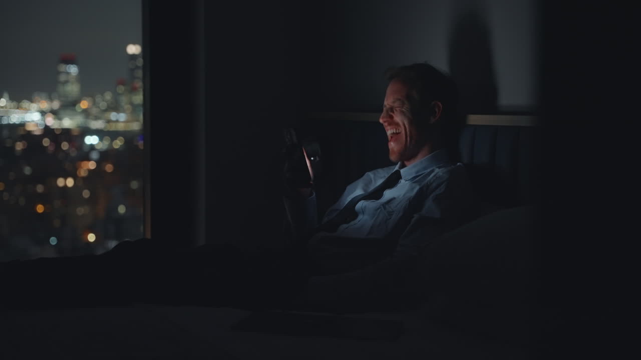 Businessman speaking to his family over video conversation while away for business trip in hotel at night overlooking the city. Apartment or house man is talking and enjoying video call sitting in bed