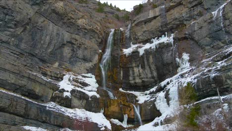 Epic-drone-scenic-waterfall-over-frozen-ice,-rocks,-cliffs,-and-snow-on-Mount-Timpanogos-in-American-Fork,-Utah-in-while-snowflakes-fall-in-foreground