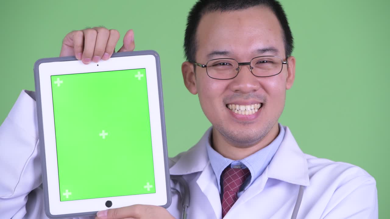 녹색 배경에 디지털 태블릿에서 복사 공간을 보여주고 미소 짓는 아시아 남성 의사의 스튜디오 초상화